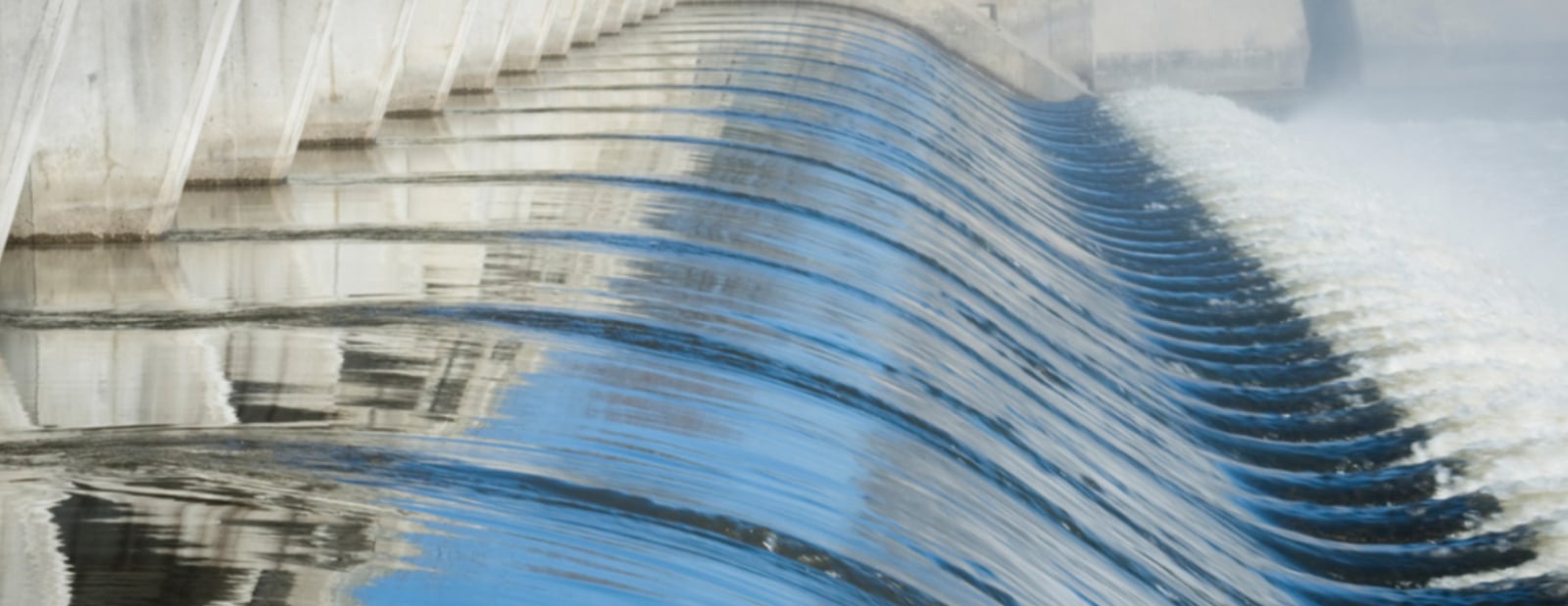 The image shows water flowing over a dam, with a smooth wave movement and reflections of the structure.