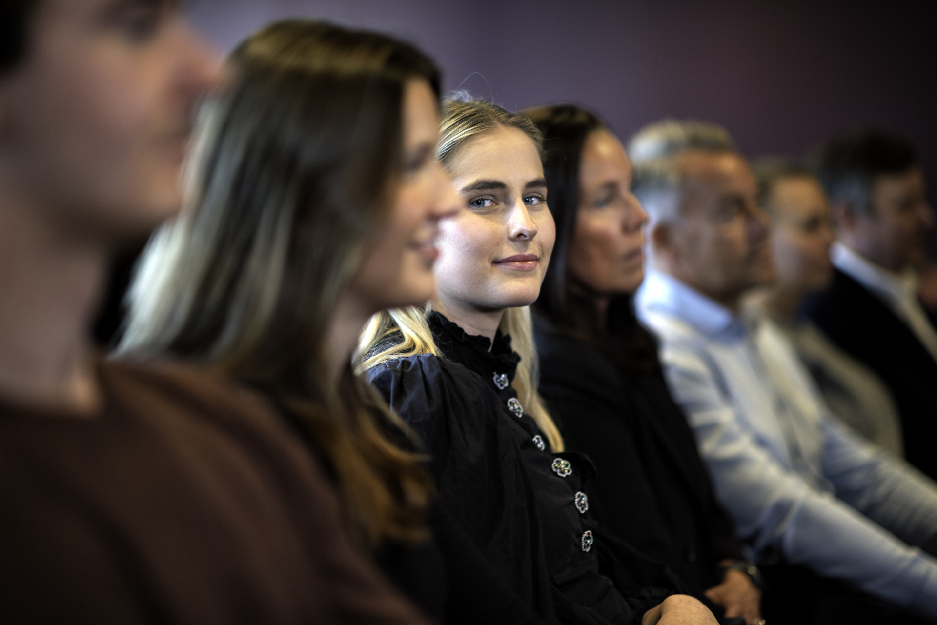 Ansatte sitter bortover på en rekke. Ung jente ser i kamera og er i fokus. 