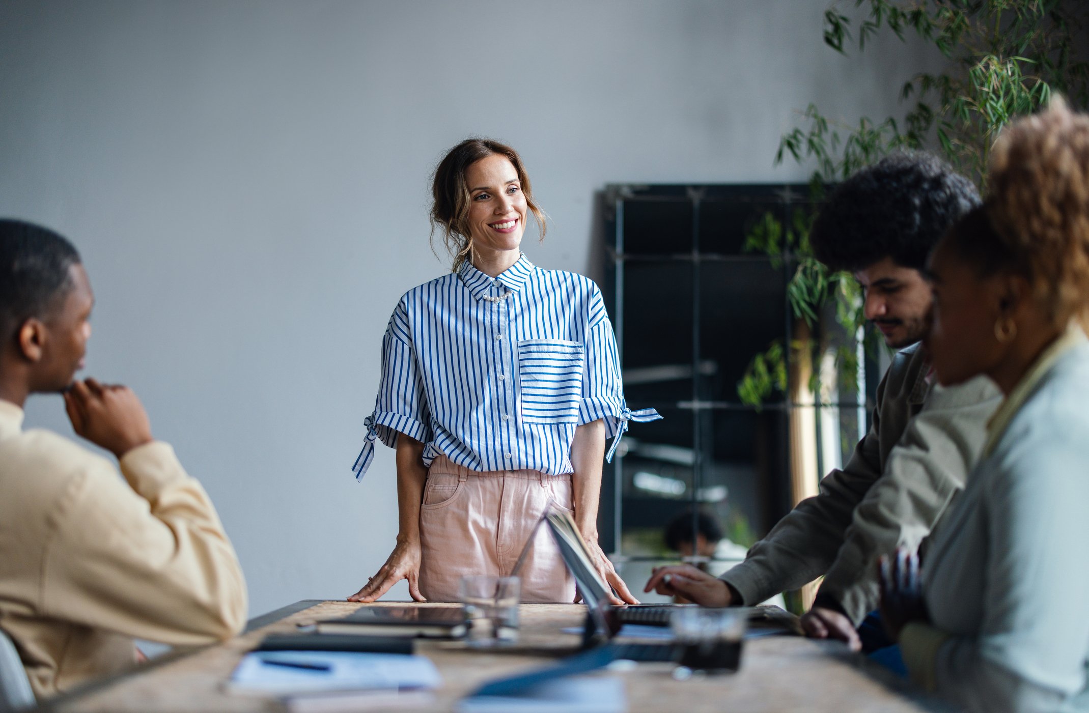A woman stands in front of a table and gives a presentation to three colleagues in a bright and modern meeting room.