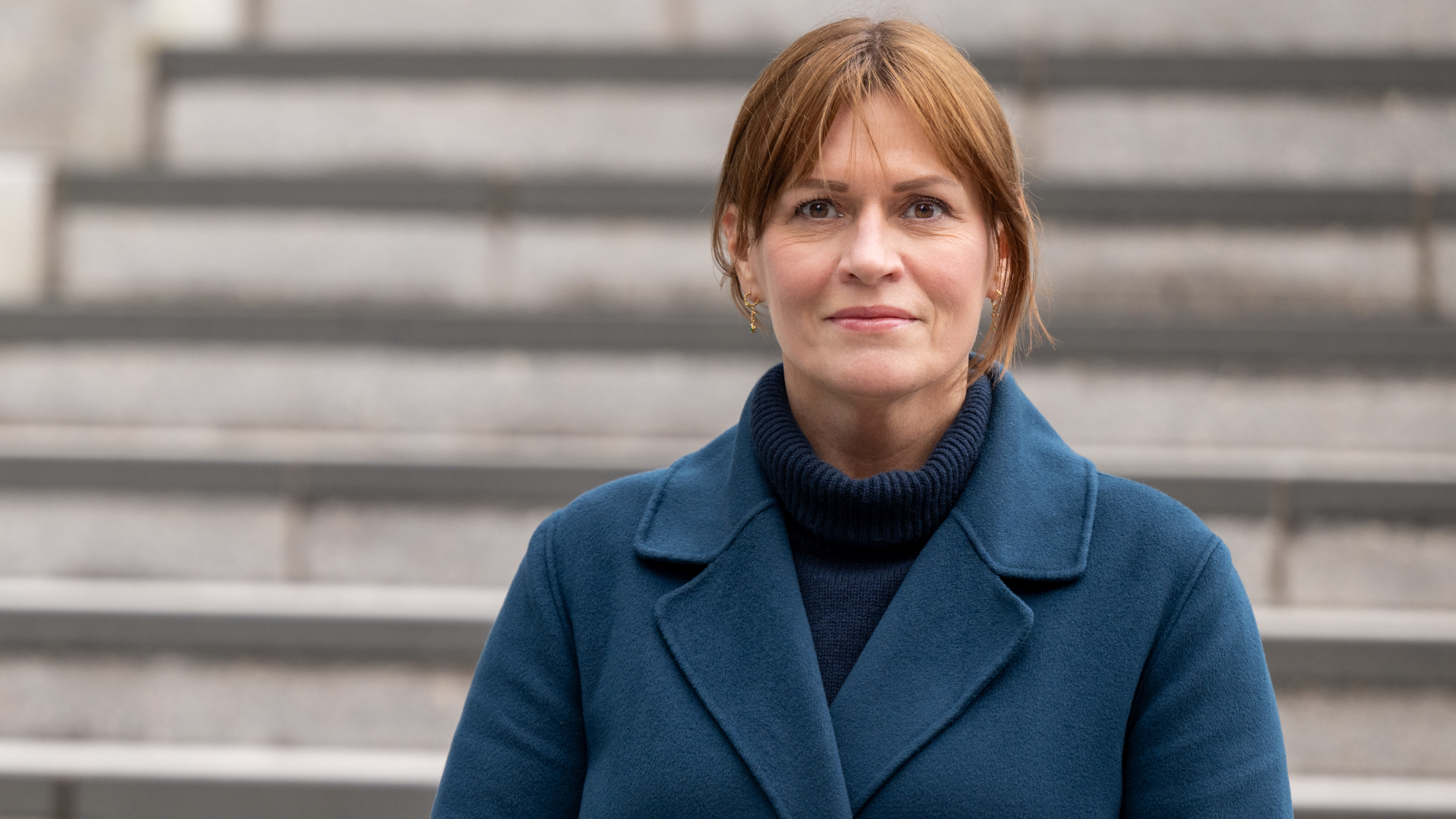 Kvinne med h&oslash;yhalset genser, bl&aring; k&aring;pe og brunt h&aring;r som ser inn i kamera