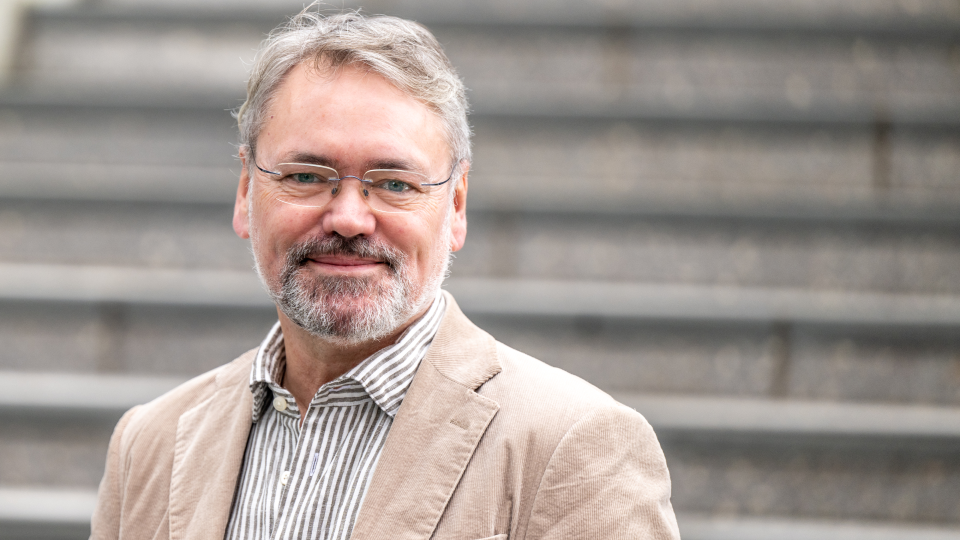 En mann med briller og gr&aring;tt skjegg, med lys blazer og stripete skjorte st&aring;r foran gr&aring; trappetrinn.