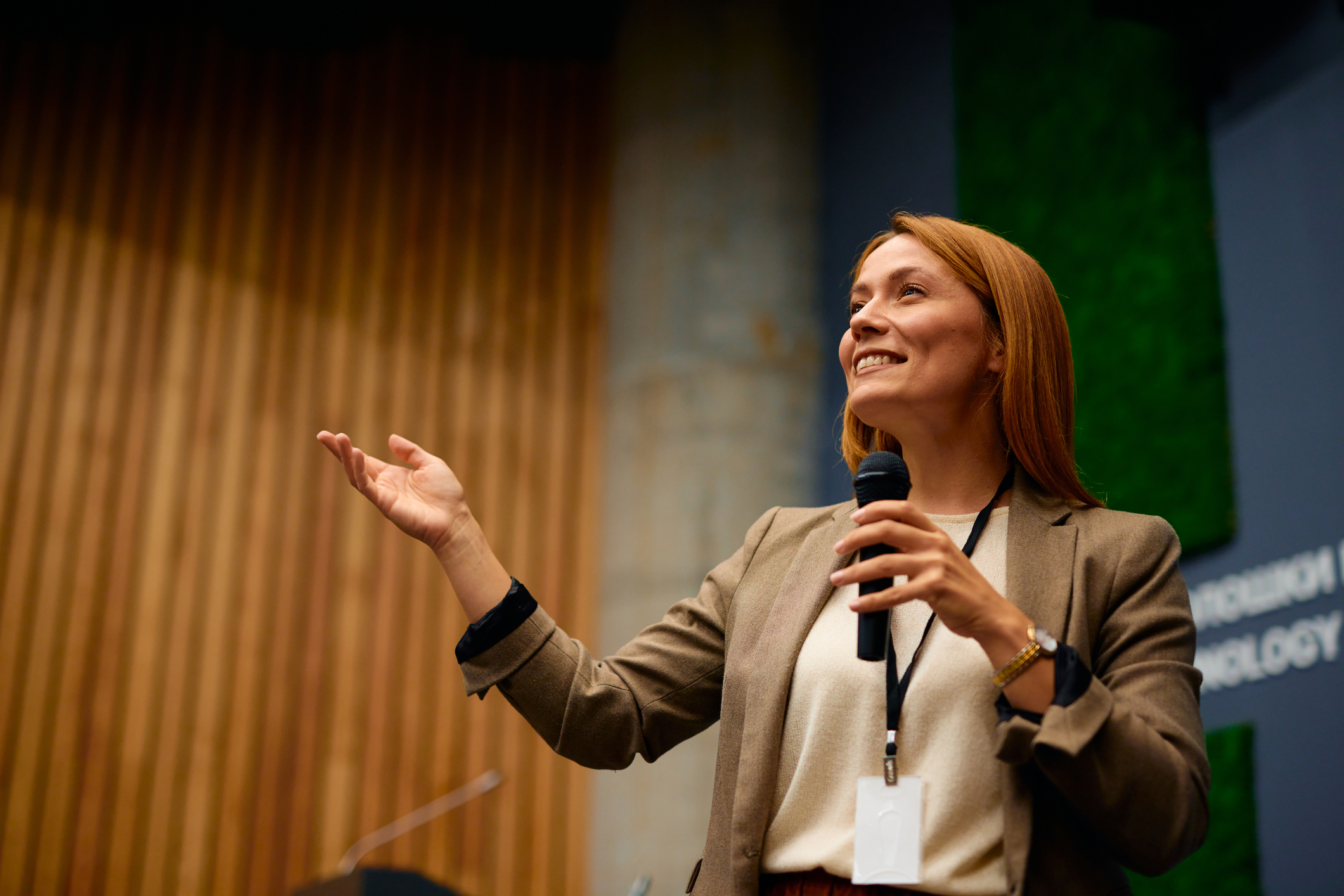 A woman with long, blond hair holds a microphone and smiles as she gestures with her hand in front of an audience in a modern conference room.