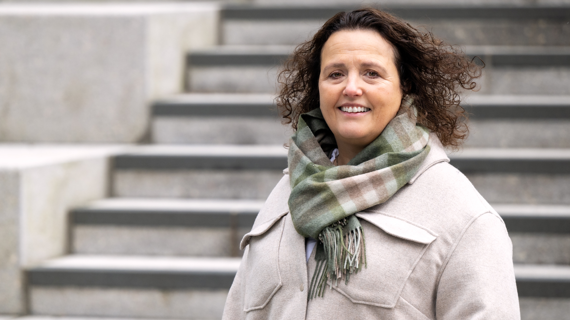 En kvinne med kr&oslash;llete h&aring;r, lys frakk og rutete skjerf st&aring;r foran gr&aring; trappetrinn.