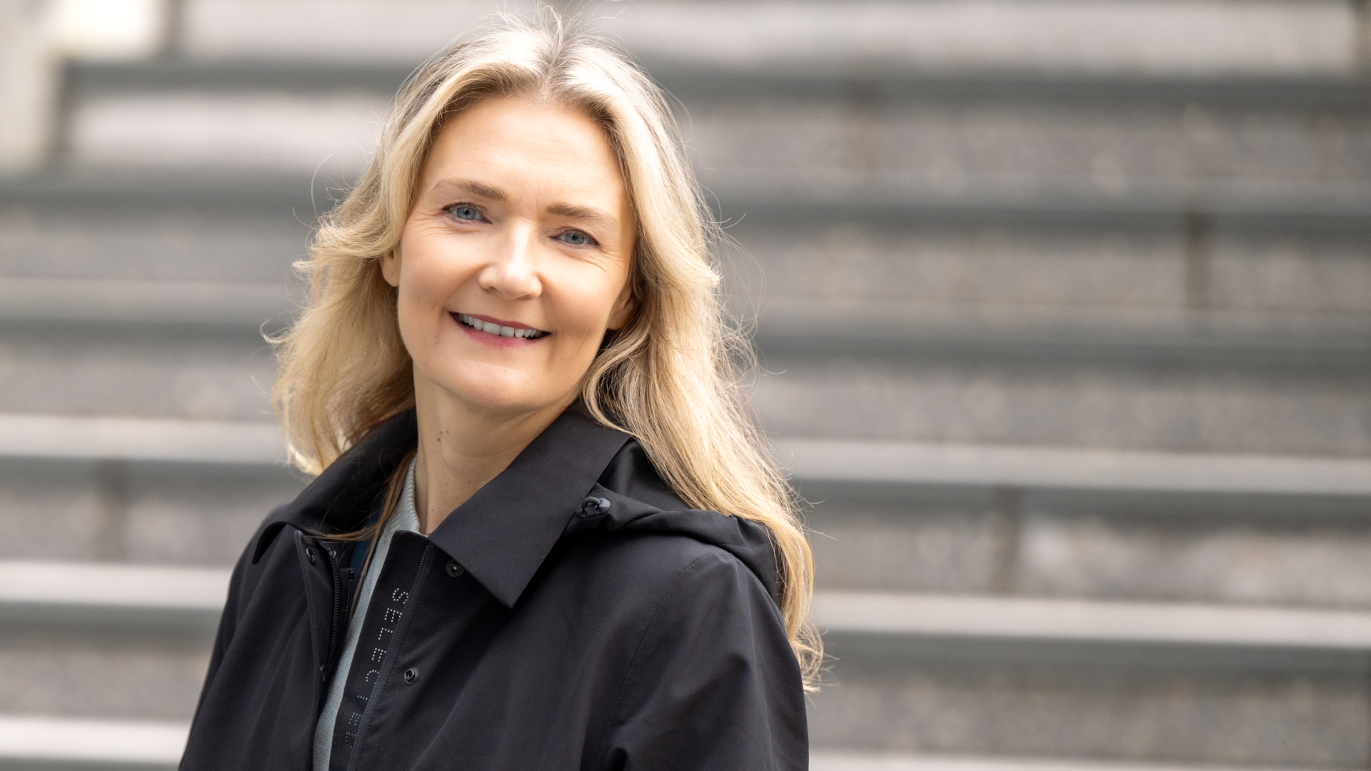 En kvinne med lyst h&aring;r if&oslash;rt m&oslash;rk jakke st&aring;r foran gr&aring; trappetrinn.