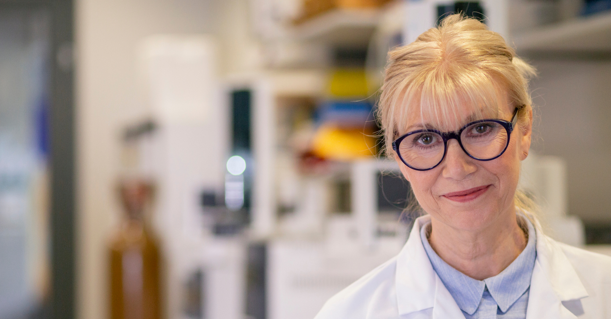 En kvinne i hvit laboratoriefrakk smiler vennlig. Hun har lyst hår, briller og står i et moderne laboratorium.