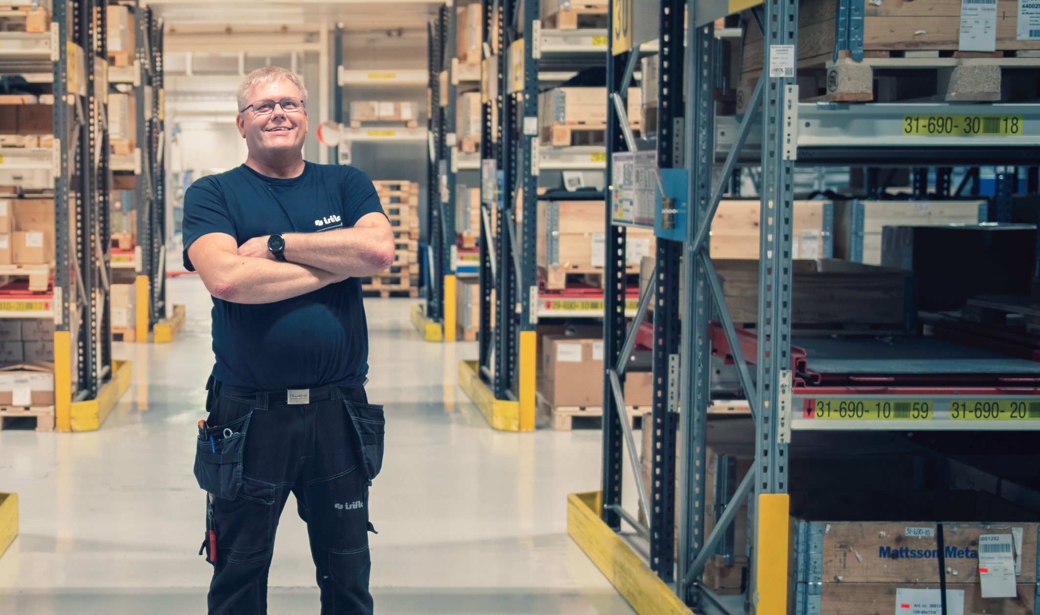 En smilende industriarbeider står med armene i kors i et ryddig lager omgitt av reoler fulle av trekasser og esker.
