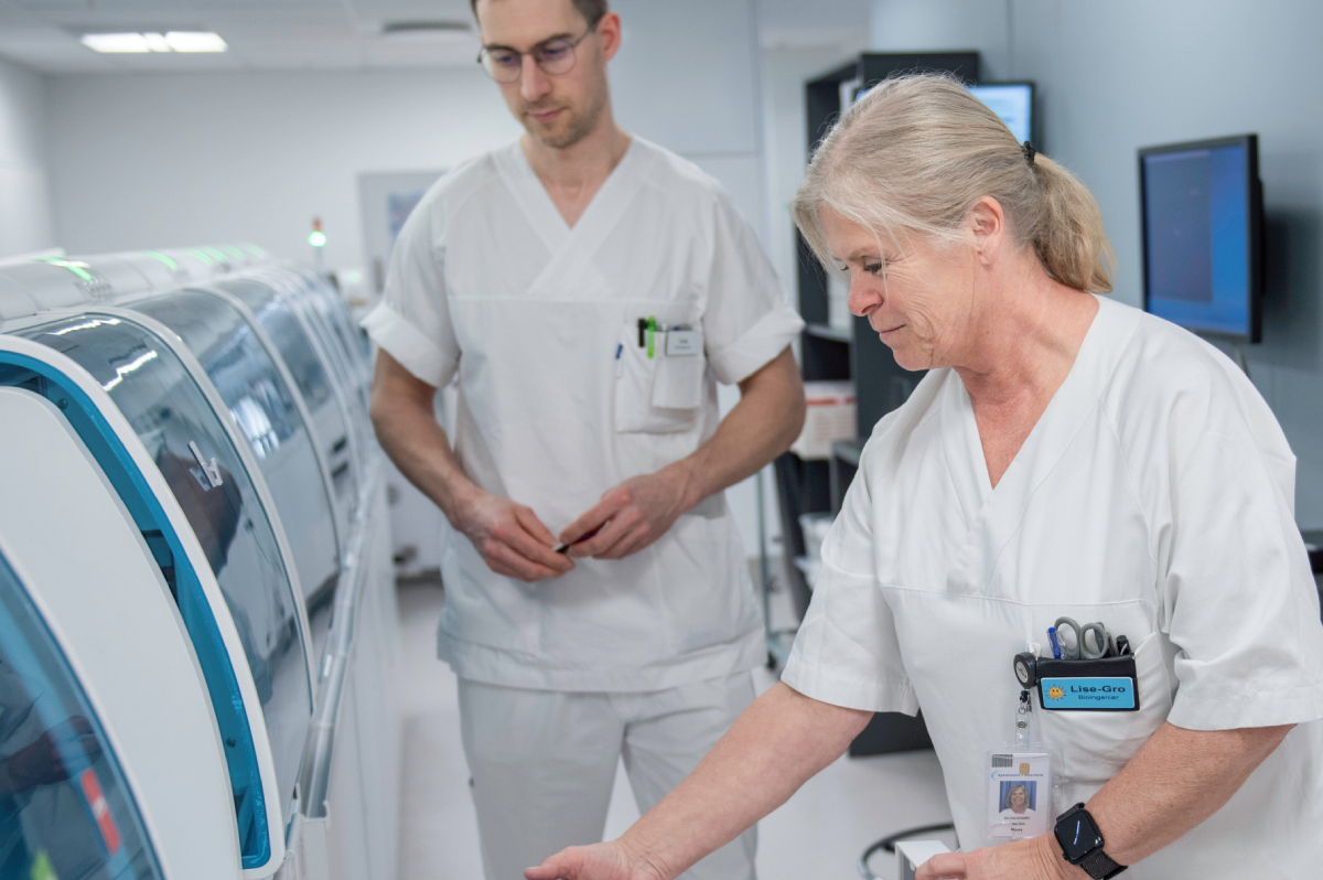 To personer i hvite laboratorieklær håndterer prøver i et moderne laboratorium, ved en stor maskin for medisinsk eller vitenskapelig analyse.
