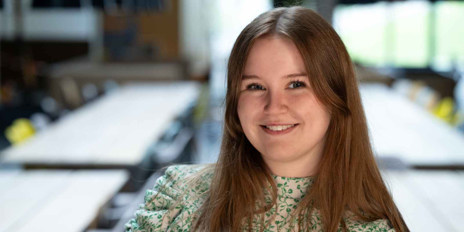 Ung kvinne med langt, brunt h&aring;r i gr&oslash;nn og hvit bluse, foran bord og stoler i et rom.