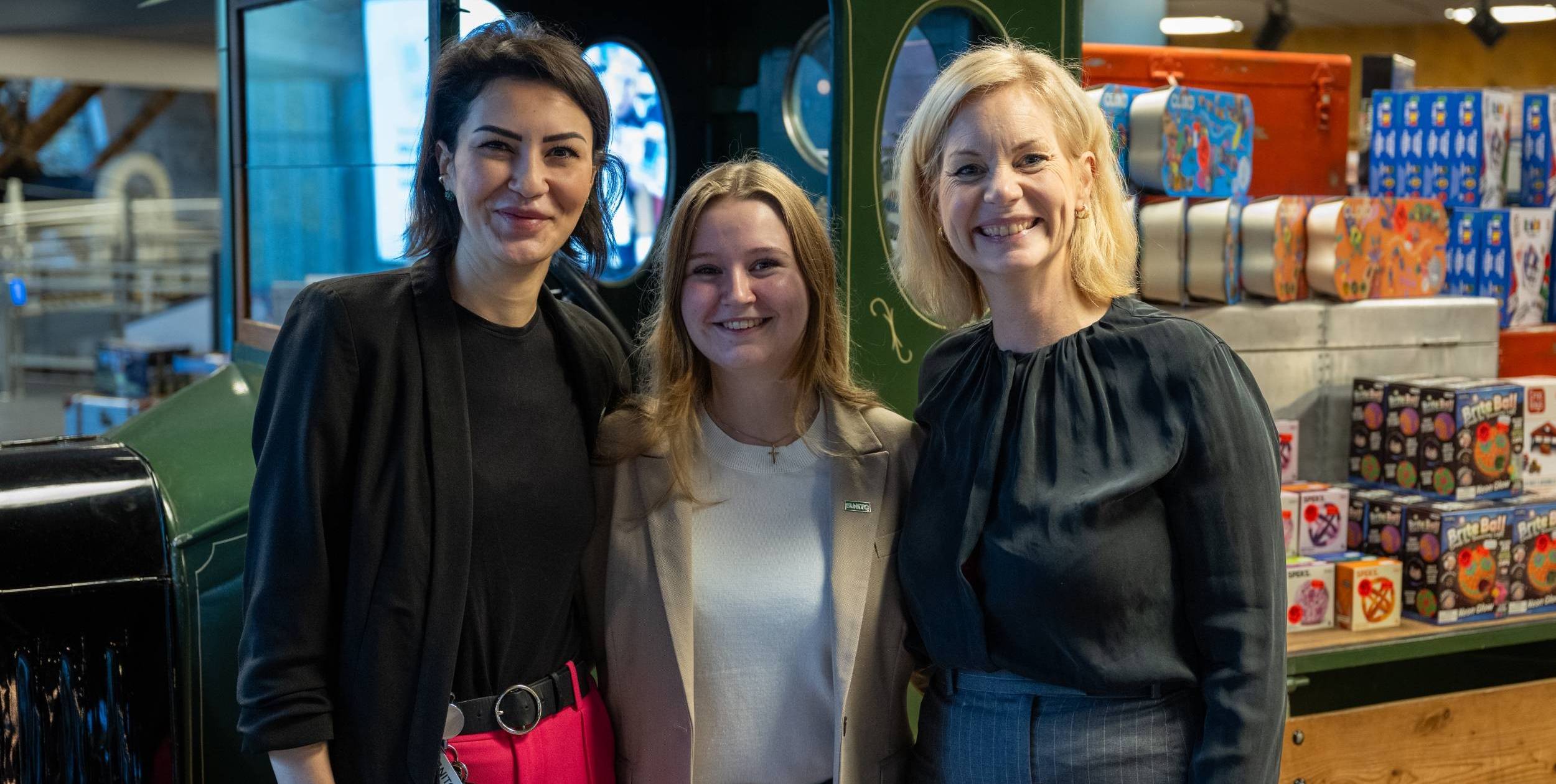 Three women stand close together and smile, surrounded by colorful boxes and industrial interiors.