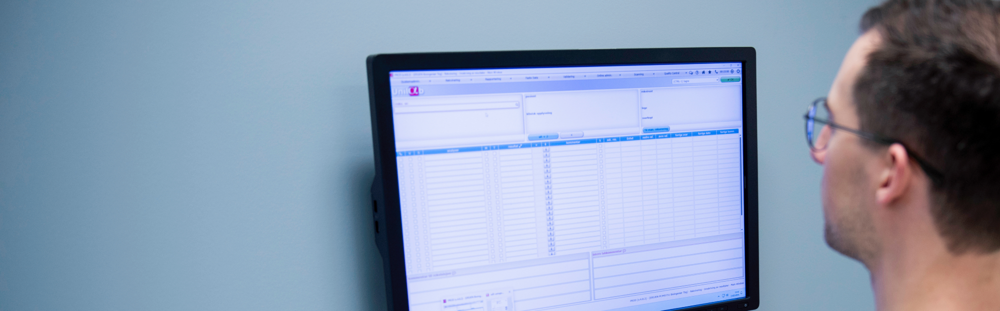 A computer screen shows a quality management system with tables and fields for laboratory data on a bright blue wall.