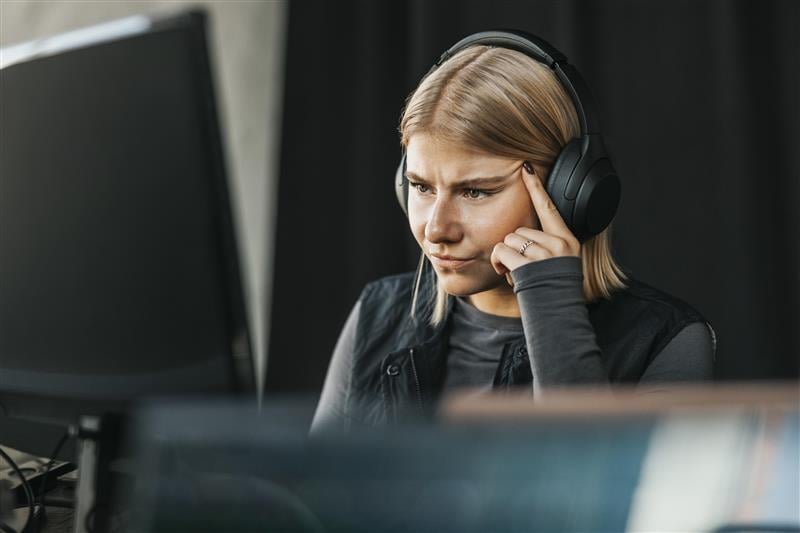 En kvinne med hodetelefoner ser konsentrert på en skjerm, med en hånd ved tinningen, som om hun tenker dypt over ord og uttrykk.