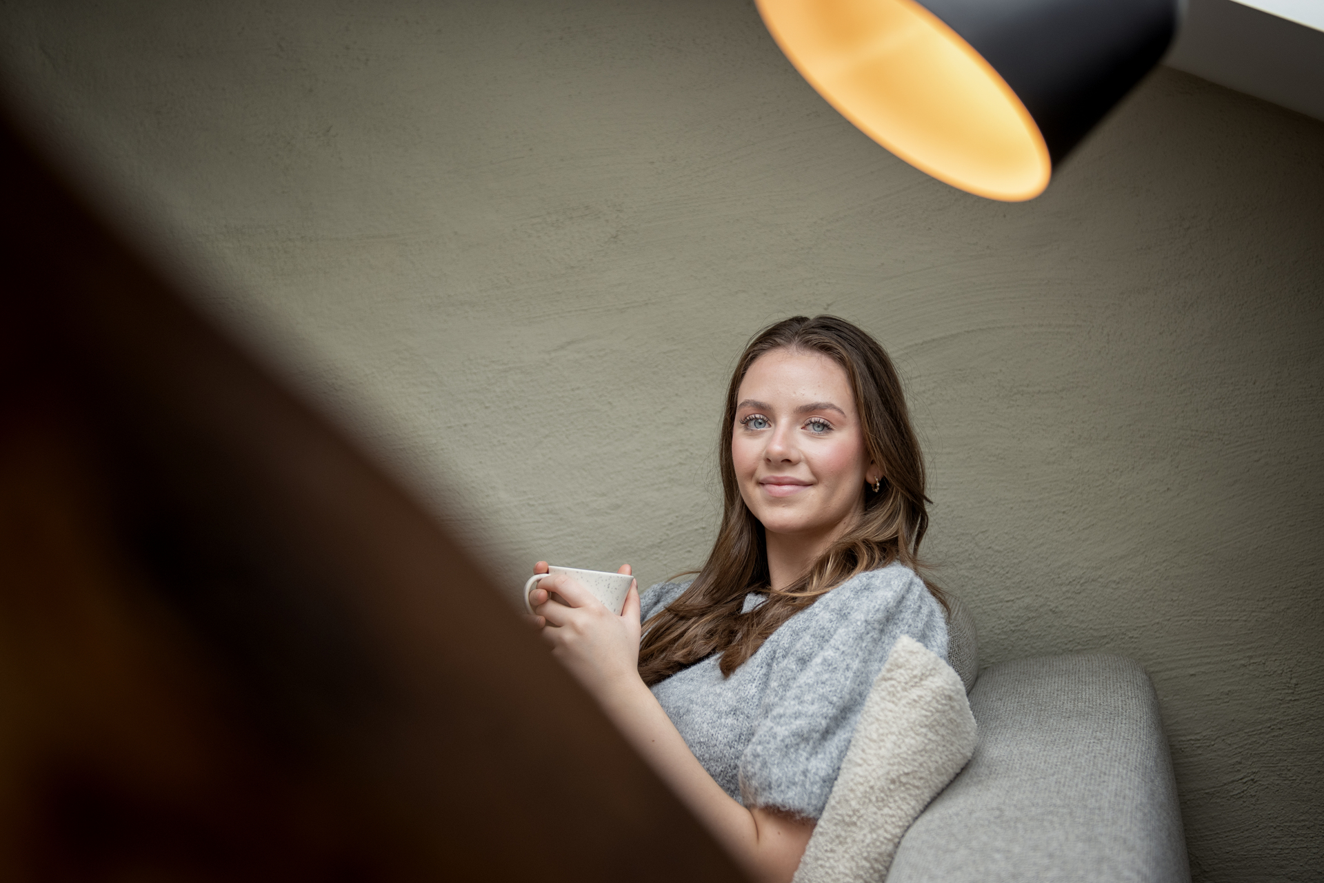Ung student sitter med kaffekopp i sofa under en lampe. 