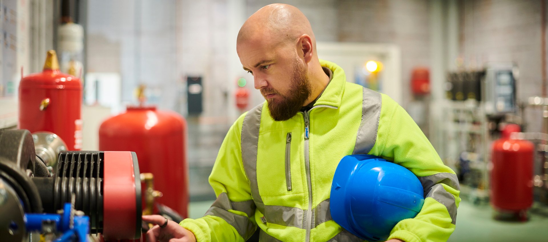 An engineer in a yellow reflective vest and a blue helmet under his arm inspects machinery in an industrial environment.