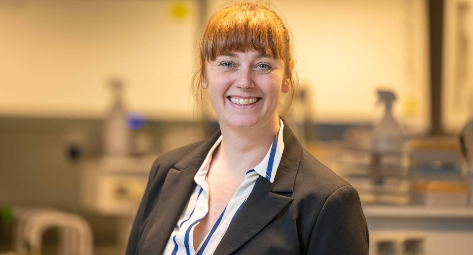 En kvinne med r&oslash;dlig h&aring;r i hestehale smiler, if&oslash;rt m&oslash;rk blazer og skjorte, st&aring;r i et lyst rom som ligner et laboratorium.