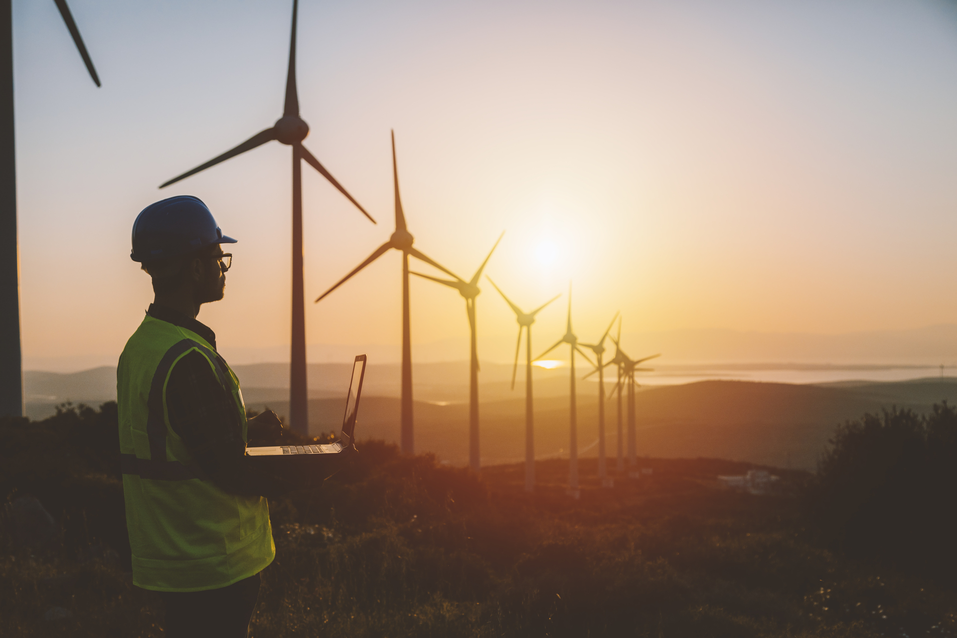 Young male engineer with laptop working standing next to windmills, symbolizing renewable energy and wage growth.