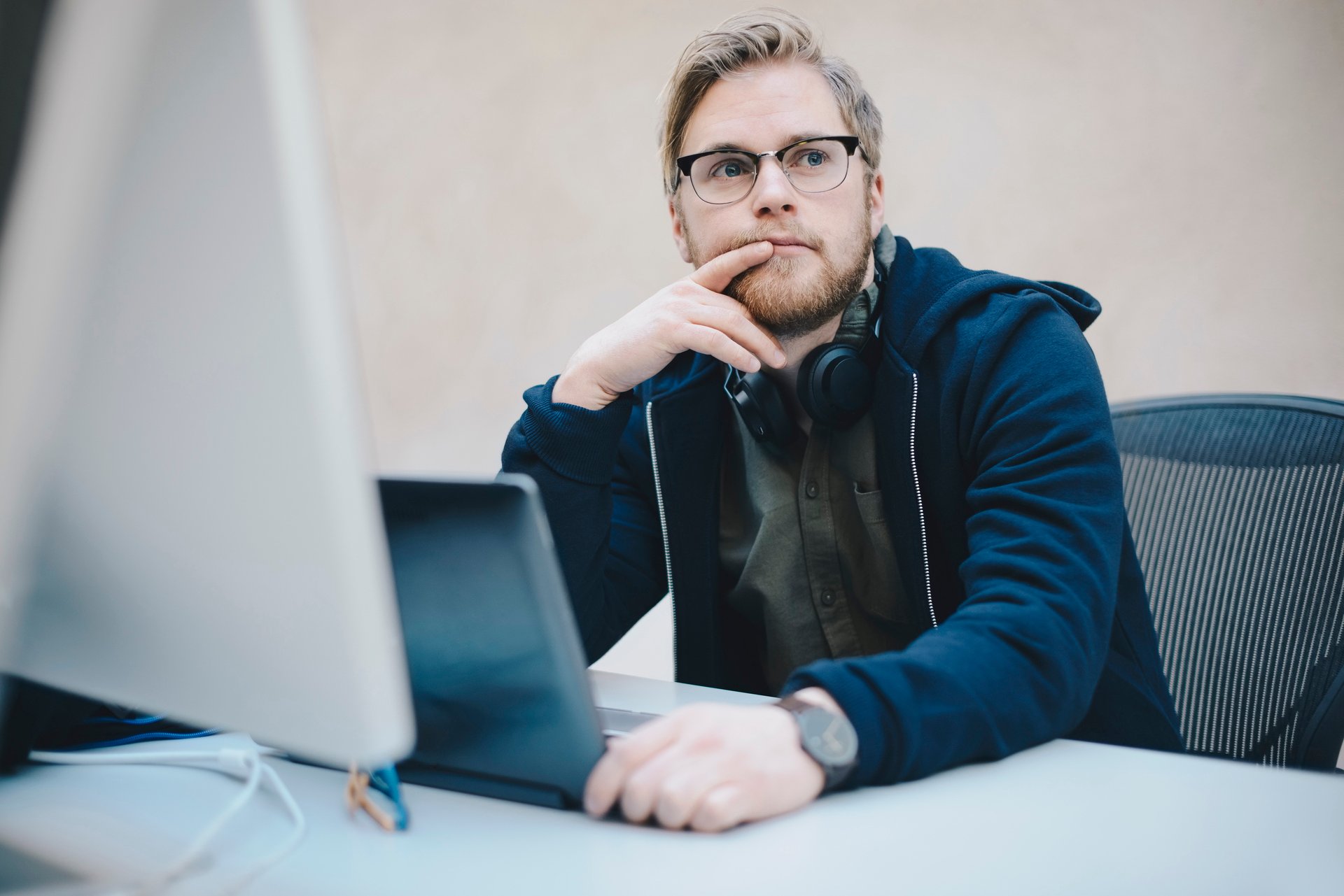 Young man sits in front of a laptop in a green sweater and looks questioningly into the air