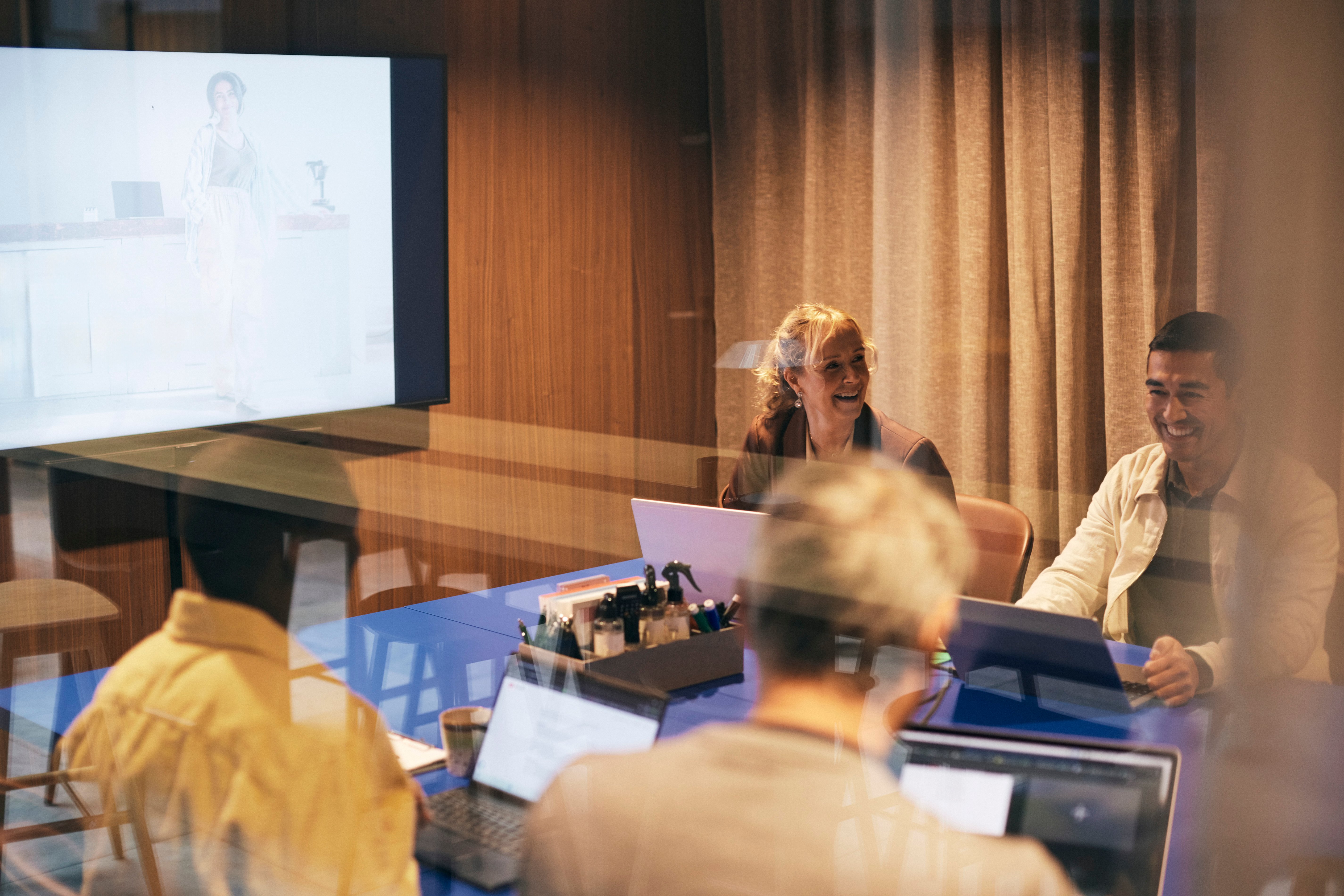 Internasjonalt samarbeidsmøte med deltakere rundt et konferansebord. En person presenterer på storskjerm mens deltakerne følger med på bærbare datamaskiner i et moderne møterom med treveggpanel. Foto: GettyImages