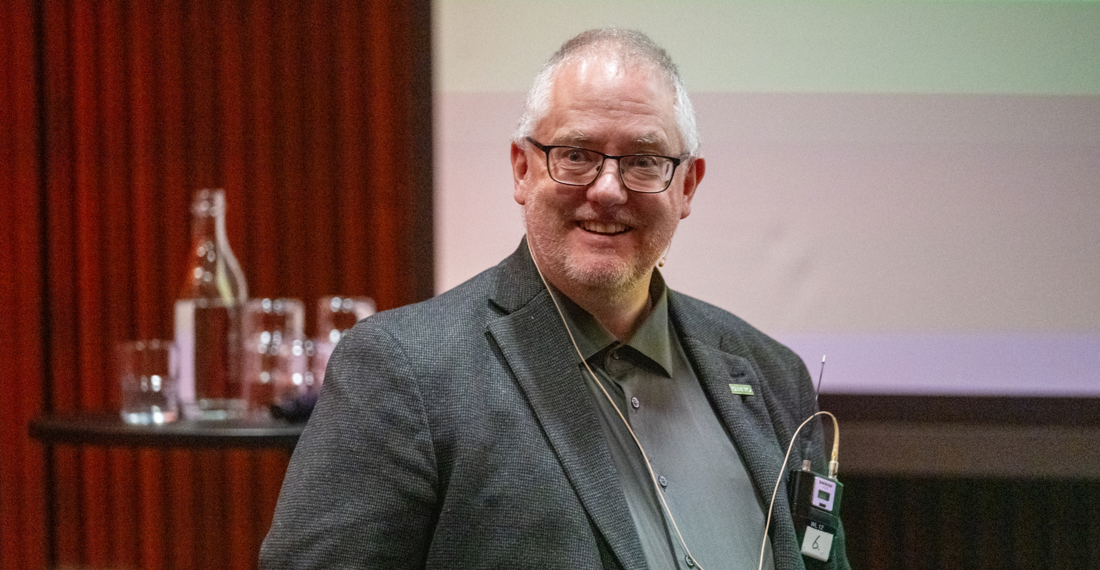 En mann i dressjakke med mikrofon st&aring;r foran publikum i et foredragssal, med bord med flasker og glass bak seg.