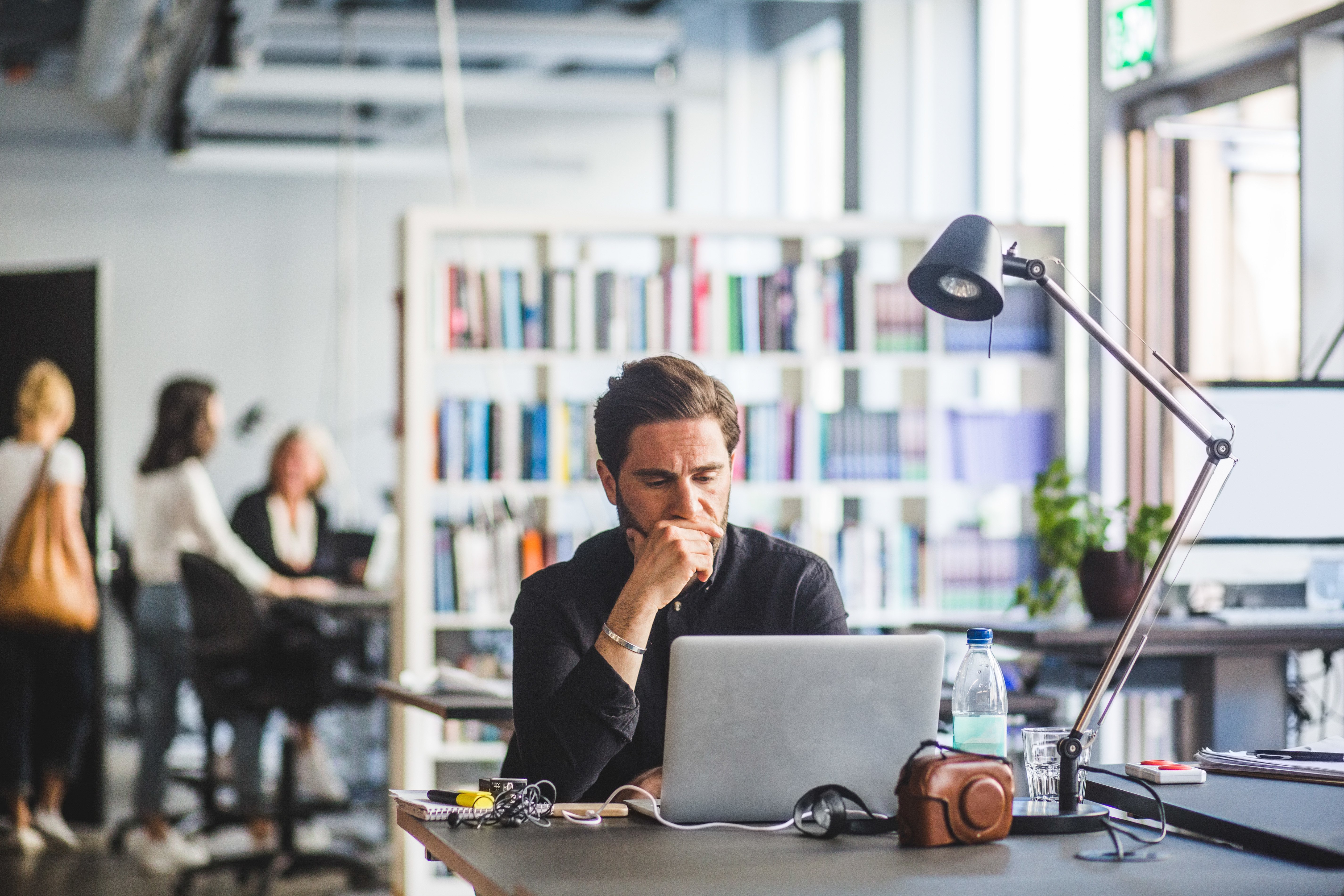 A man sits concentrated in front of a laptop in an office, with his hand on his chin, and seems to be reflecting or solving a problem. In the background, there are blurred colleagues and a bookshelf, indicating a dynamic work environment.