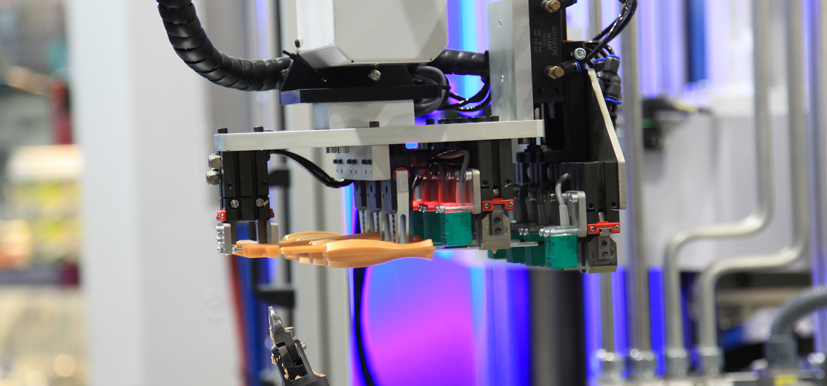 En industrirobot med gripeverktøy holder en plastdel, omgitt av kabler, metall og lilla bakgrunnslys.