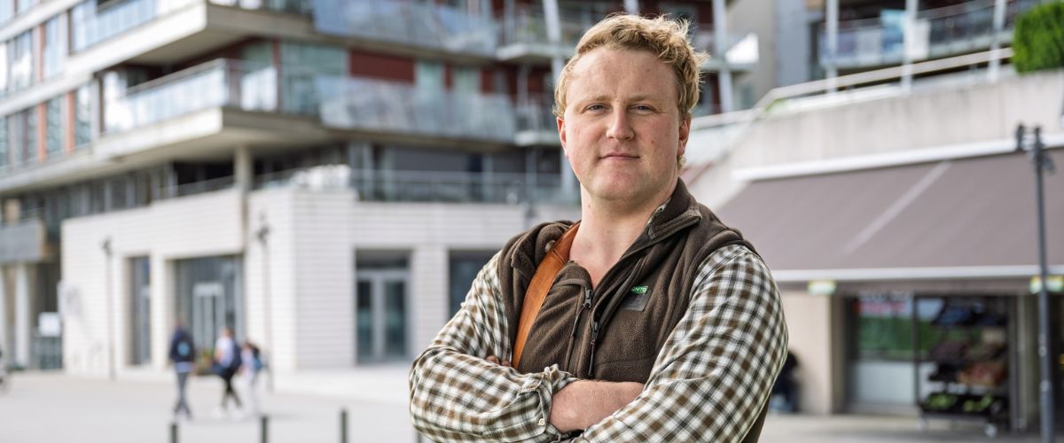 Jaradn Byrkjeland i rutete skjorte og vest står med armer i kors foran bygninger. 