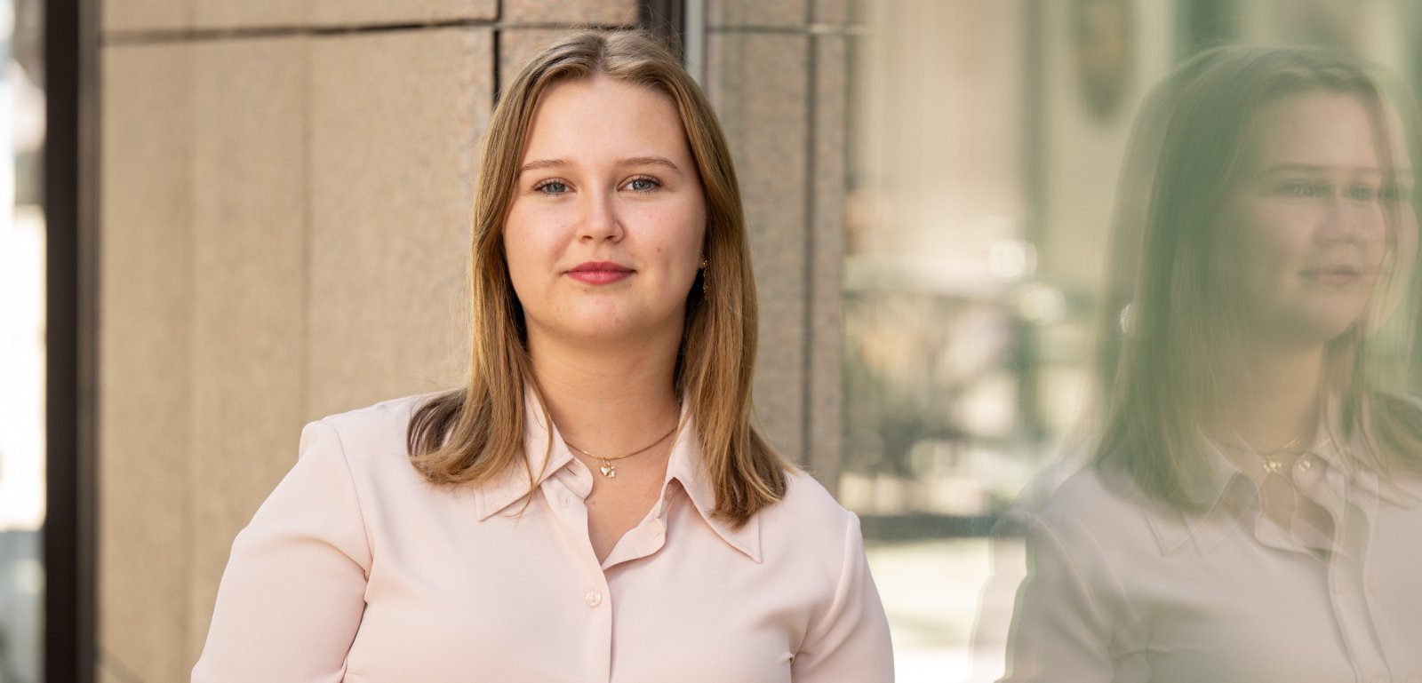 Ung kvinne med lyst hår i lys bluse står ved murvegg. Hun ser rolig inn i kameraet, med refleksjon i et vindu ved siden av.