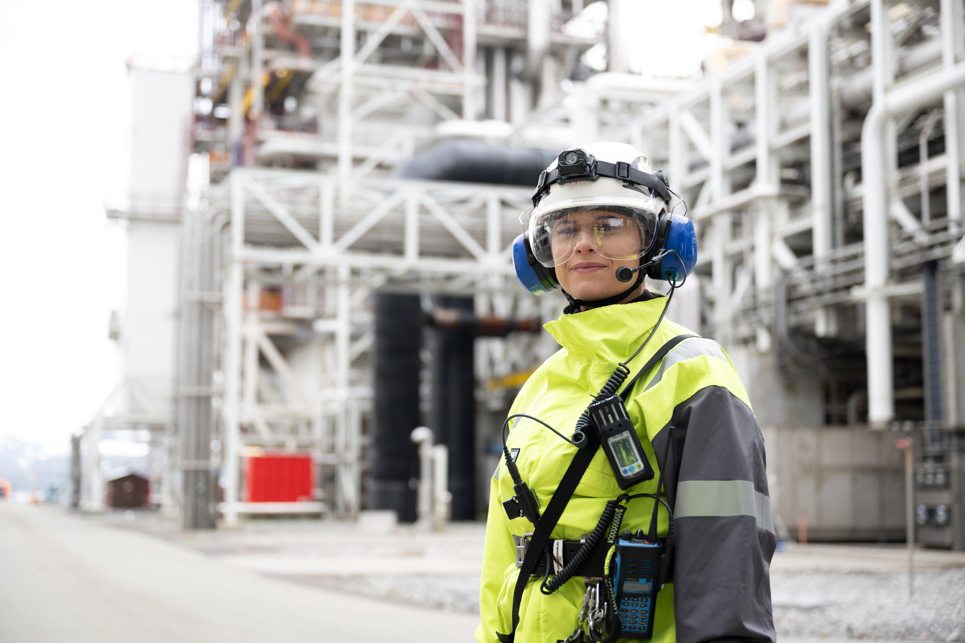 Ingeniør i neongrønn arbeidsjakke, hjelm og hørselvern står foran et industrianlegg