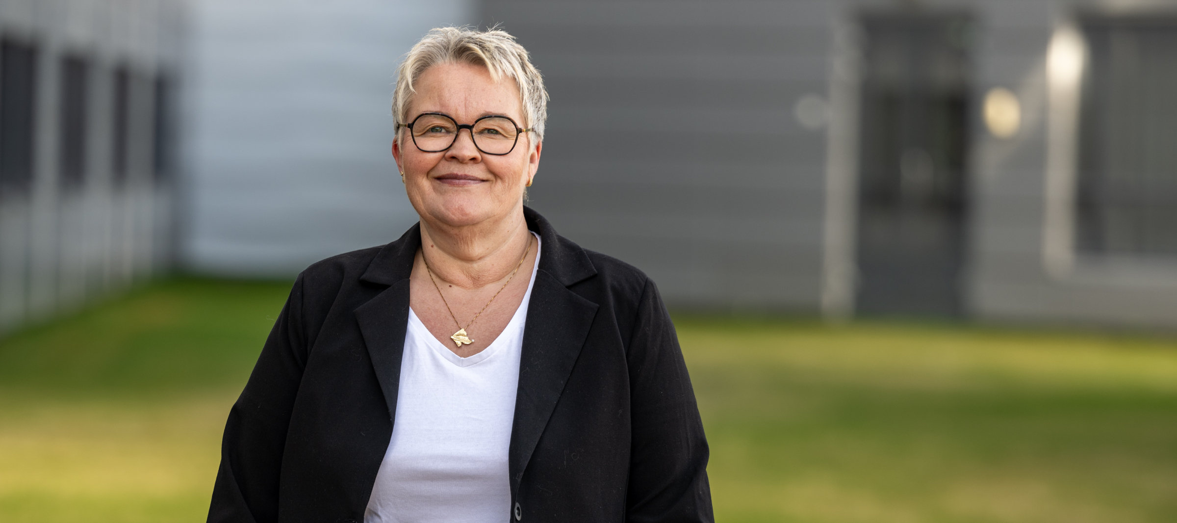 En smilende kvinne med kort, lyst hår, briller og svart jakke står utendørs foran en bygning med grønt gress i bakgrunnen.