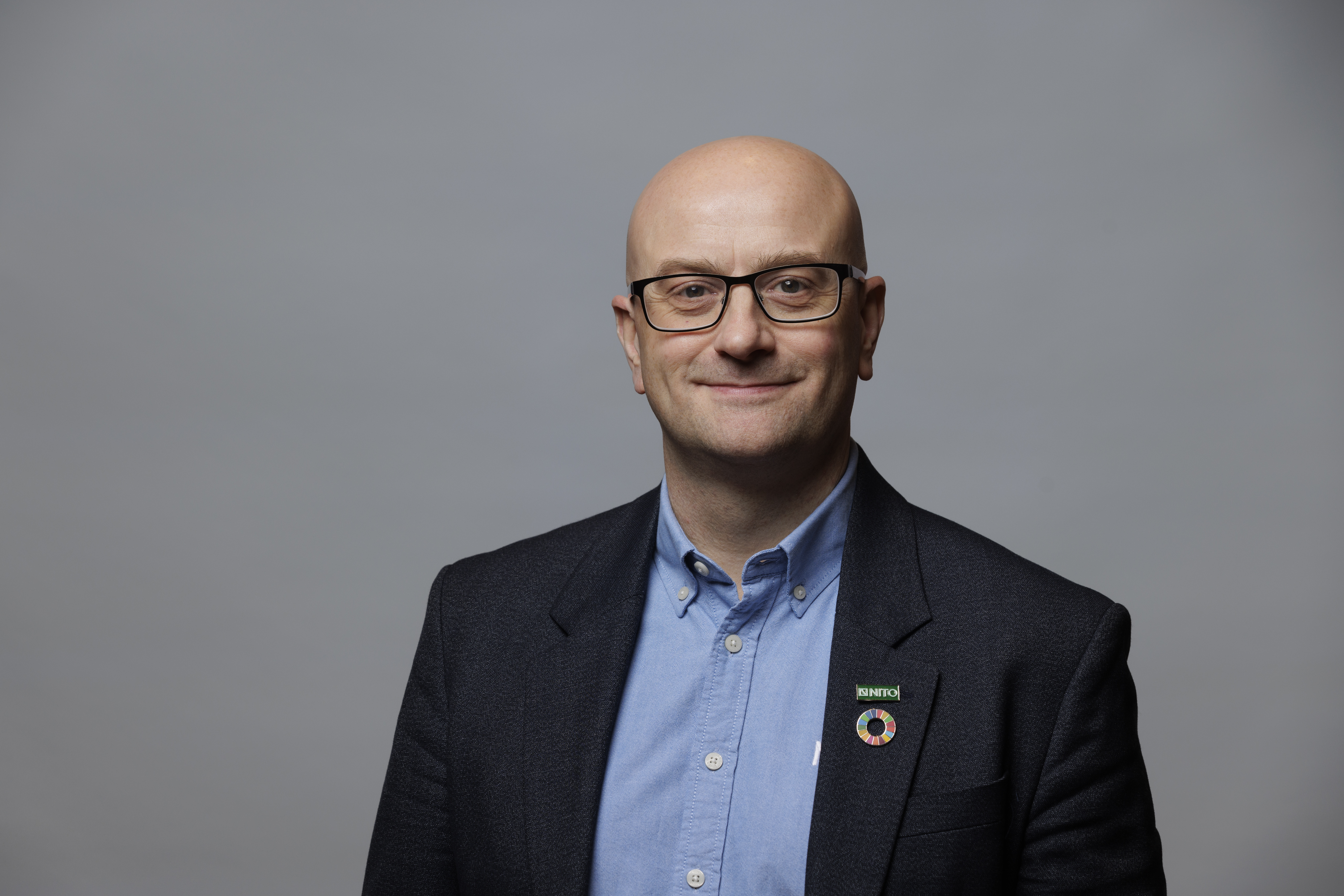 Portrait of a smiling man with glasses in a suit and shirt against a neutral grey background.