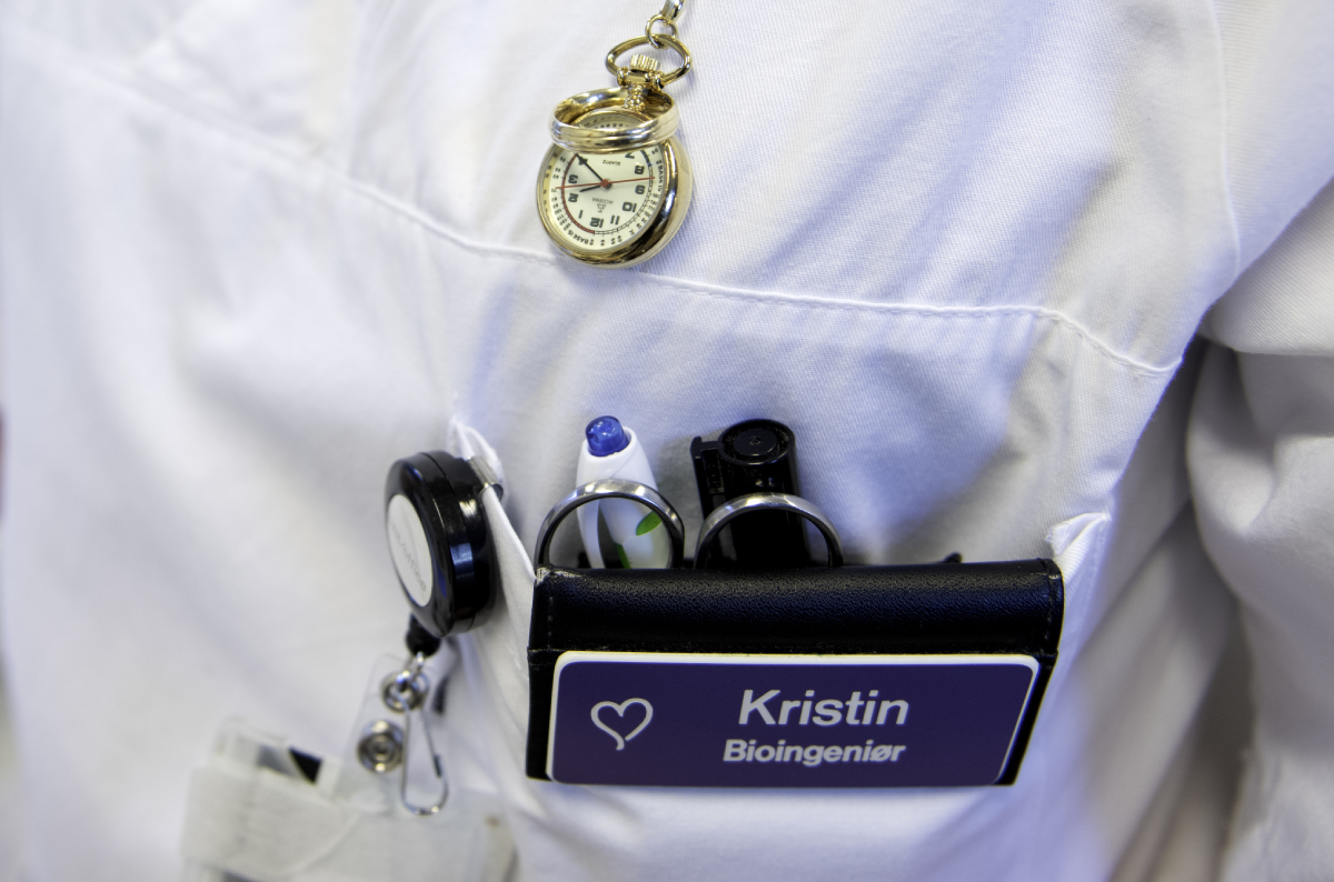 Bildet viser overkroppen til en person i hvit uniform. I brystlommen er det penner, ID-kort og et navneskilt med teksten: "Kristin, Bioingeniør". 