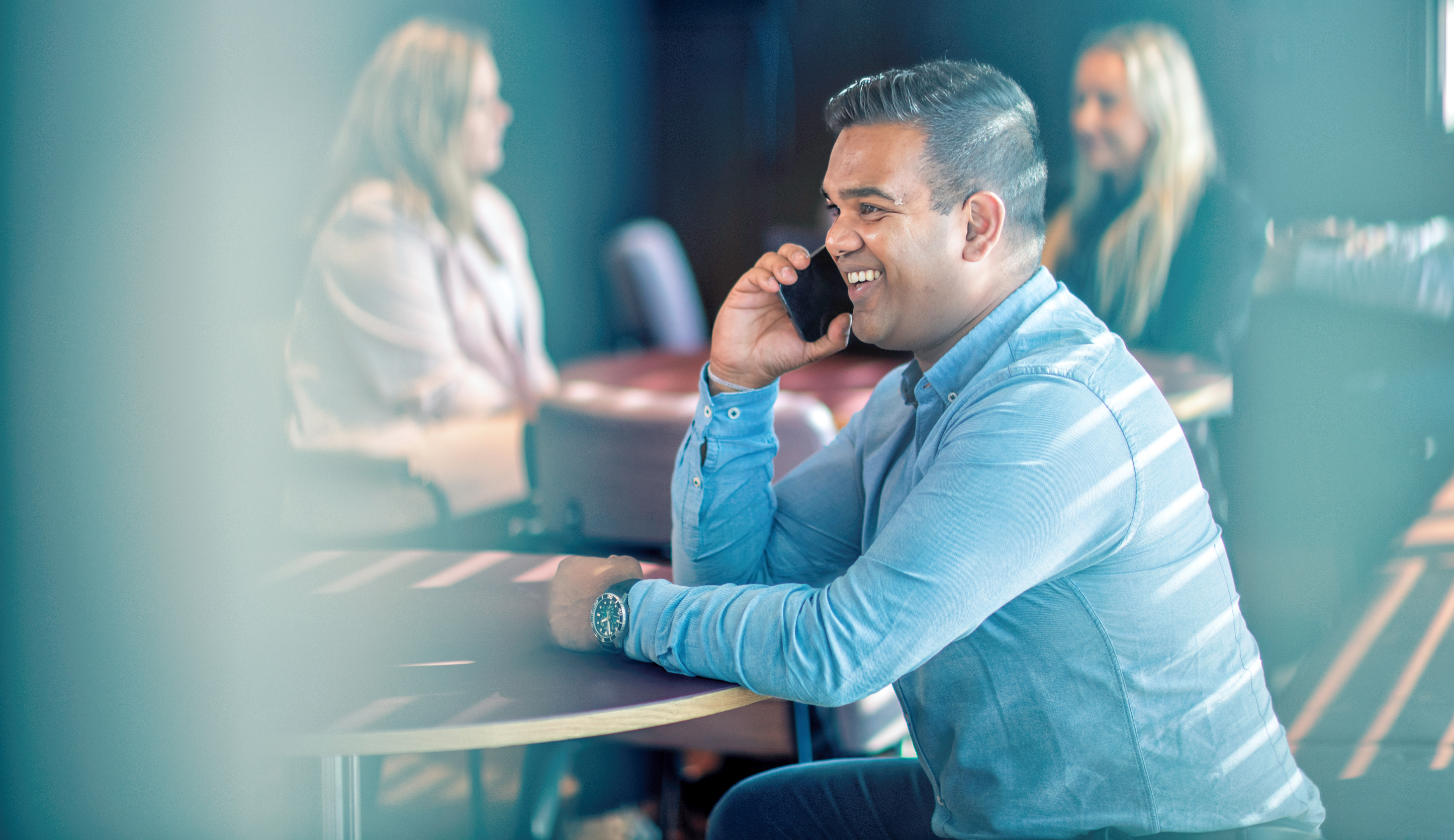 Smilende ung mann i blå skjorte som snakker i telefonen. Mennesker i bakgrunnen.