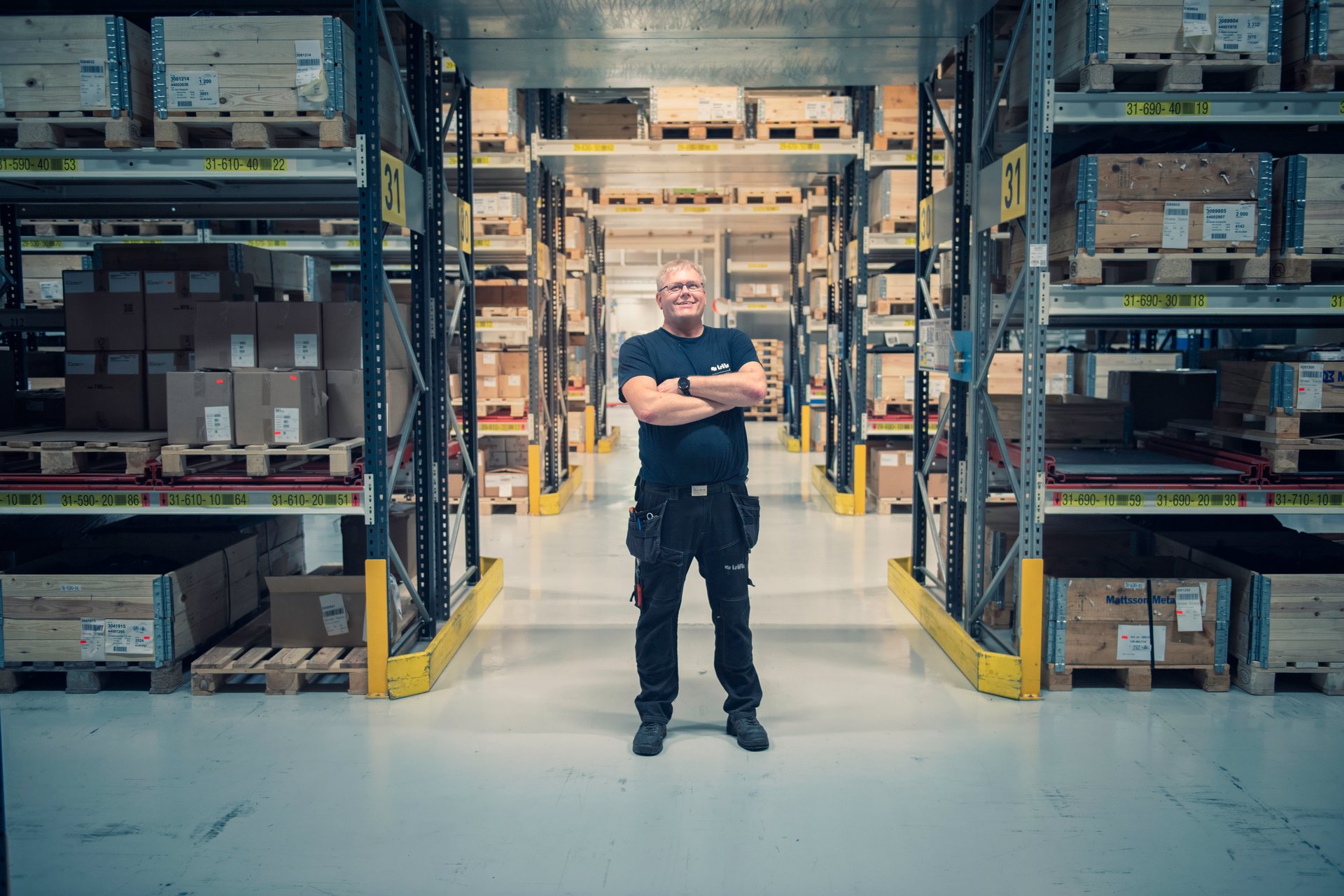  En mannlig ingeniør står i en lagerbygning omgitt av hyller med varer og smiler. Bildet illustrerer en typisk arbeidsplass hvor tjenestepensjon kan være en viktig del av de ansattes fremtidige økonomi.