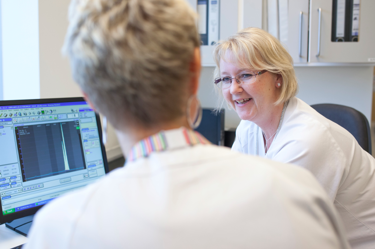 Bildet viser to bioingeni&oslash;rer i hvite frakker ved en dataskjerm. Den ene smiler og ser p&aring; den andre, som jobber ved skjermen.