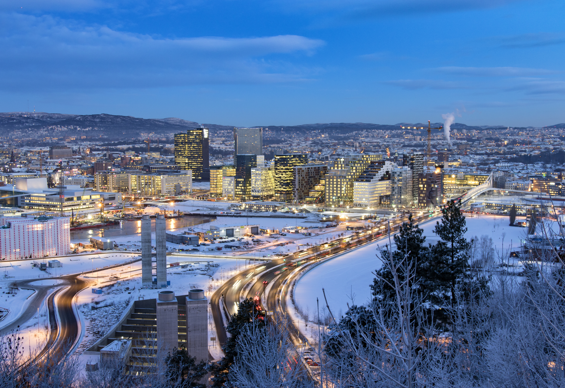 A view of Oslo city centre on a winter's evening, with snow-covered streets, illuminated buildings and blue skies in the background.