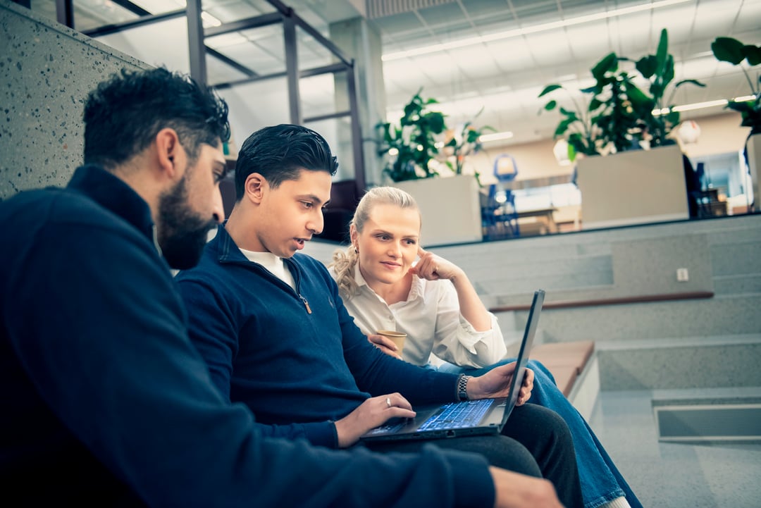 Three people are sitting together on a staircase indoors, looking at a laptop and smiling.