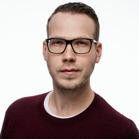 Mann med briller og lett skjegg, iført burgunderrød genser. Ser rett mot kamera. Bakgrunnen er hvit. 