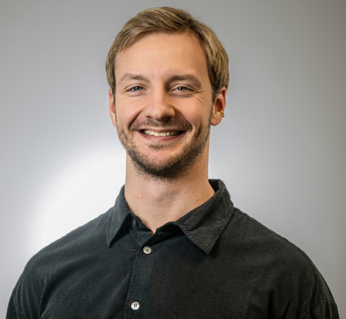 Mann med lyst hår gredd til siden og lett skjegg, iført mørk skjorte. Bakgrunnen er lys grå.