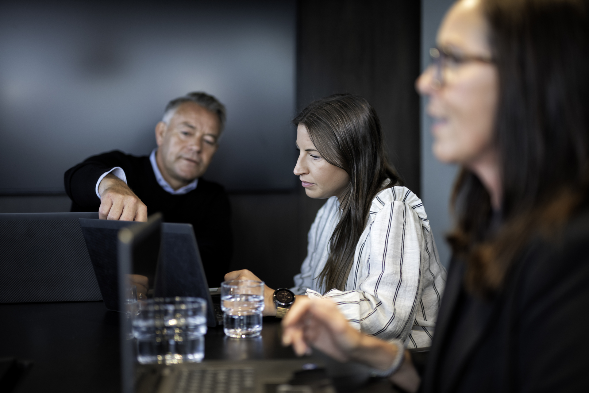 Tre ansatte i et møterom, en av dem peker på en PC-skjerm for å forklare noe til kollegaen sin. Symboliserer medbestemmelse og kommunikasjon mellom ledelsen og de ansatte.