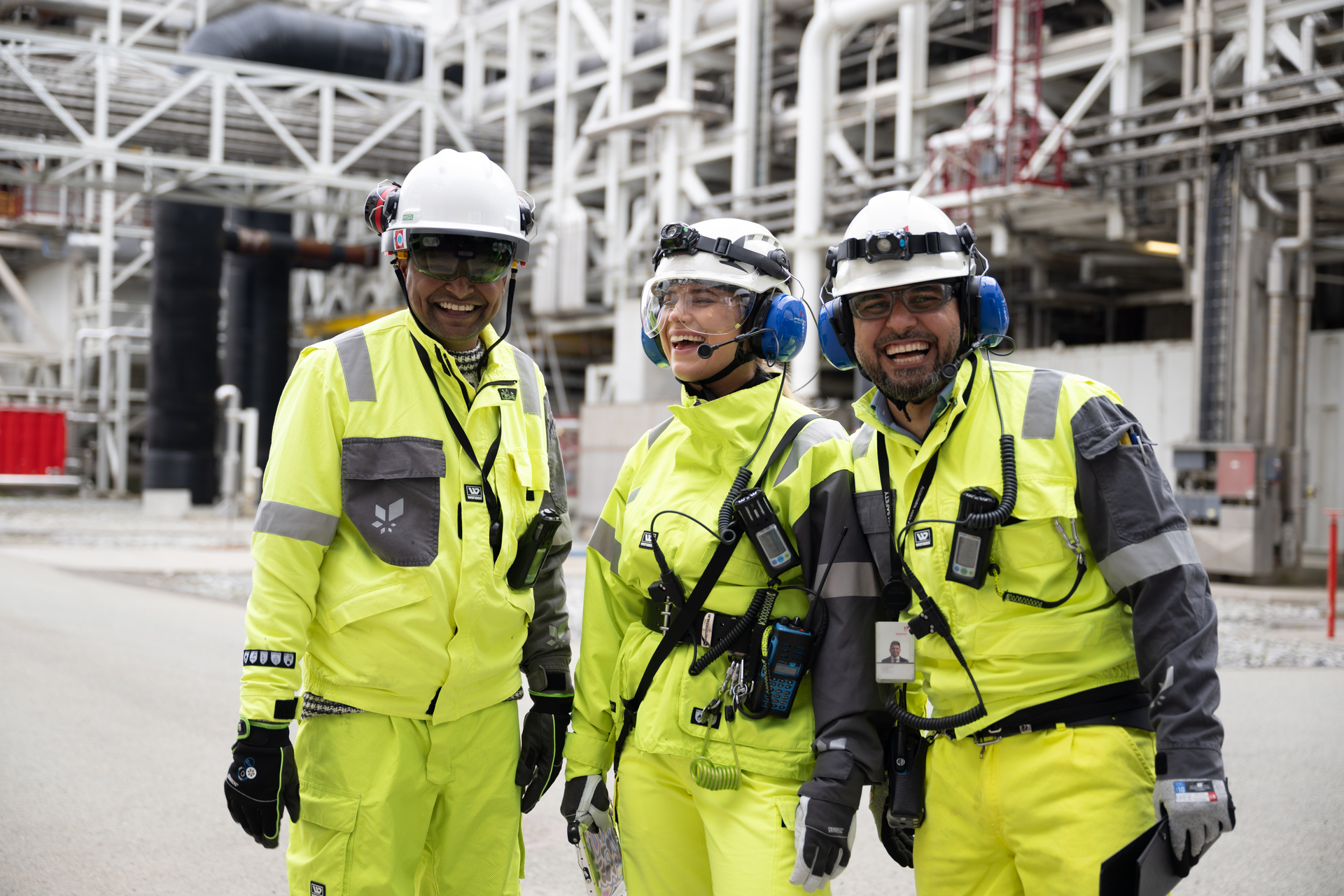 Tre ingeniører, iført verneutstyr med hjelm, smiler til kamera mens de står i et industriområde.