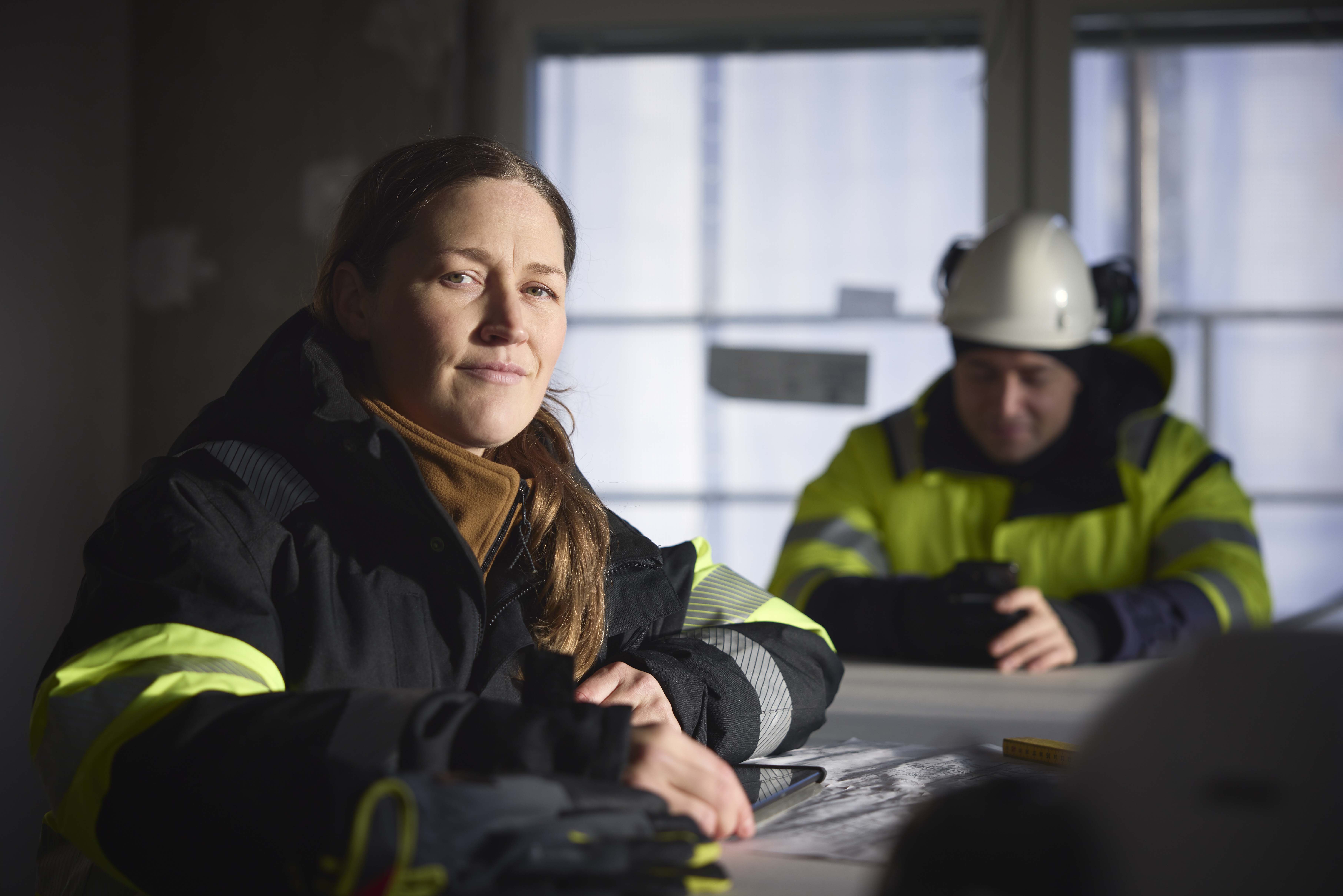 To ingeniører i refleksvest og hjelm sitter ved et bord i et mørkt rom, kvinnen smiler mot kamera mens mannen jobber på en ipad.