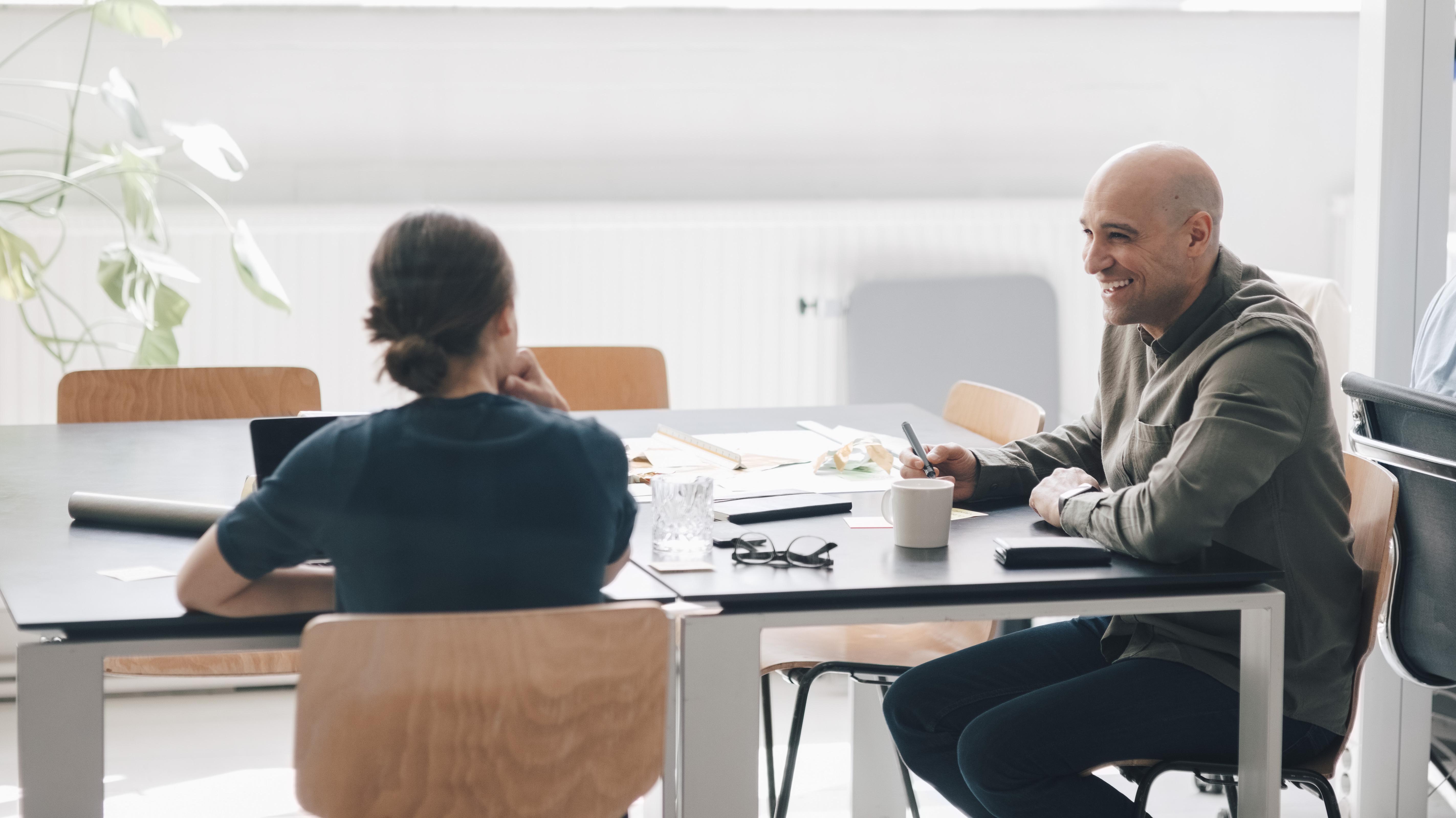Male and female colleagues in conversation in a modern office environment, the man smiles. Illustrates the other ways you can increase your salary outside of the annual negotiations.
