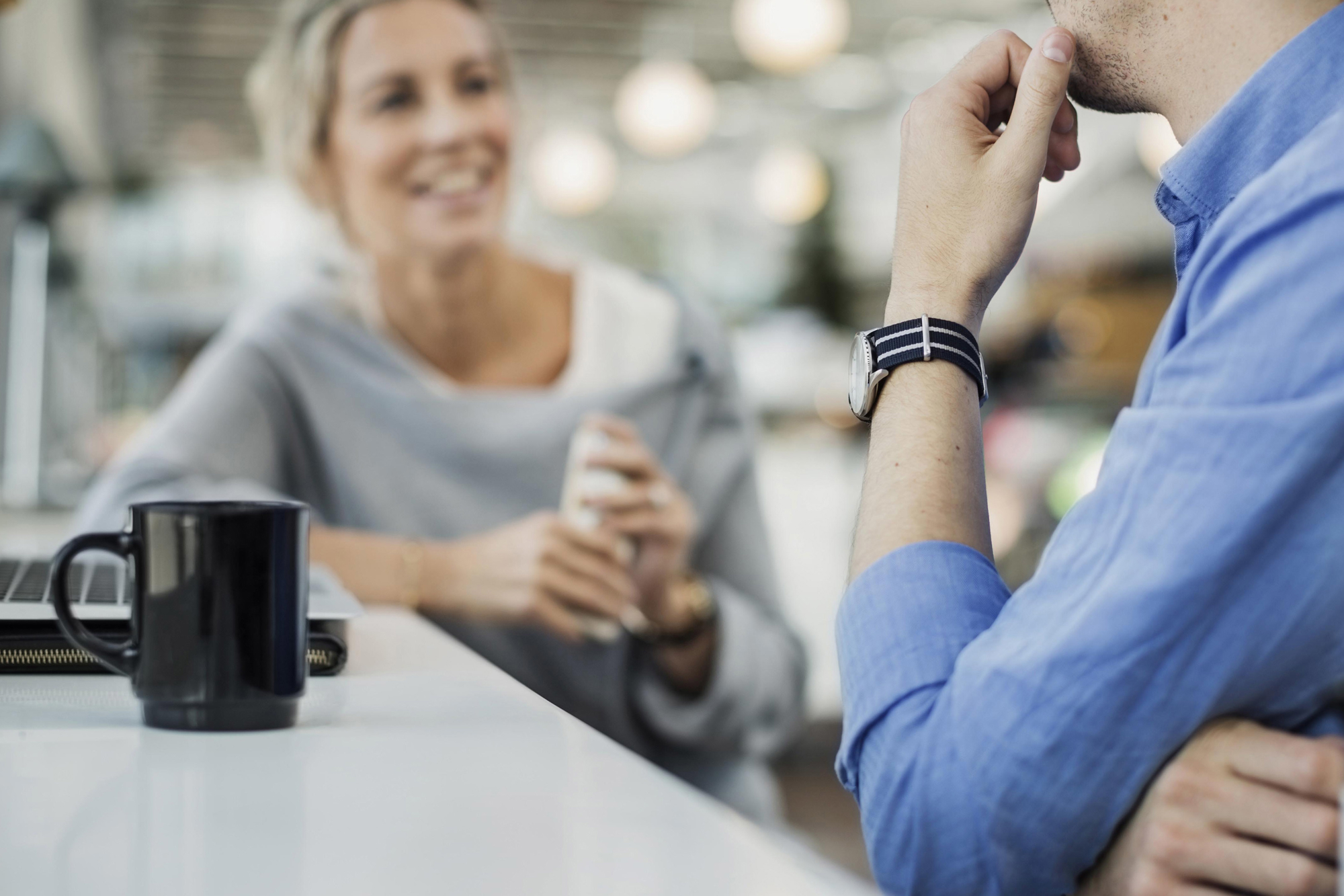 Mannlig og kvinnelig kollega i samtale over kaffe, kvinnen smiler. Illustrerer en positiv lønns- eller forhandlingssamtale.