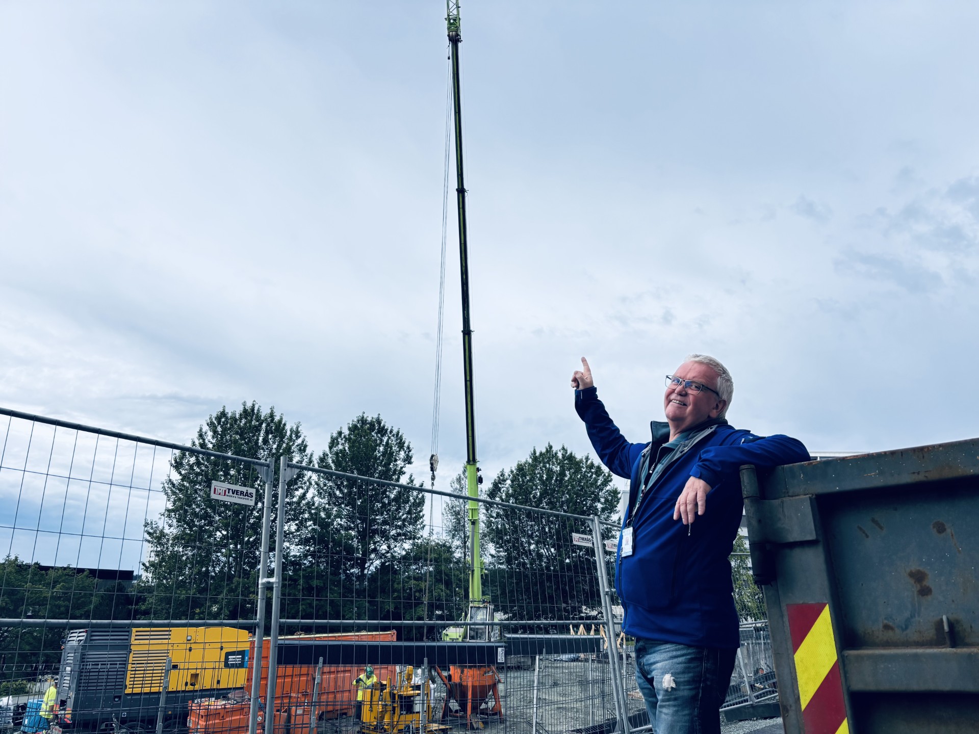 A man points up at a crane on a construction site, surrounded by trees and construction machinery under a cloudy sky.